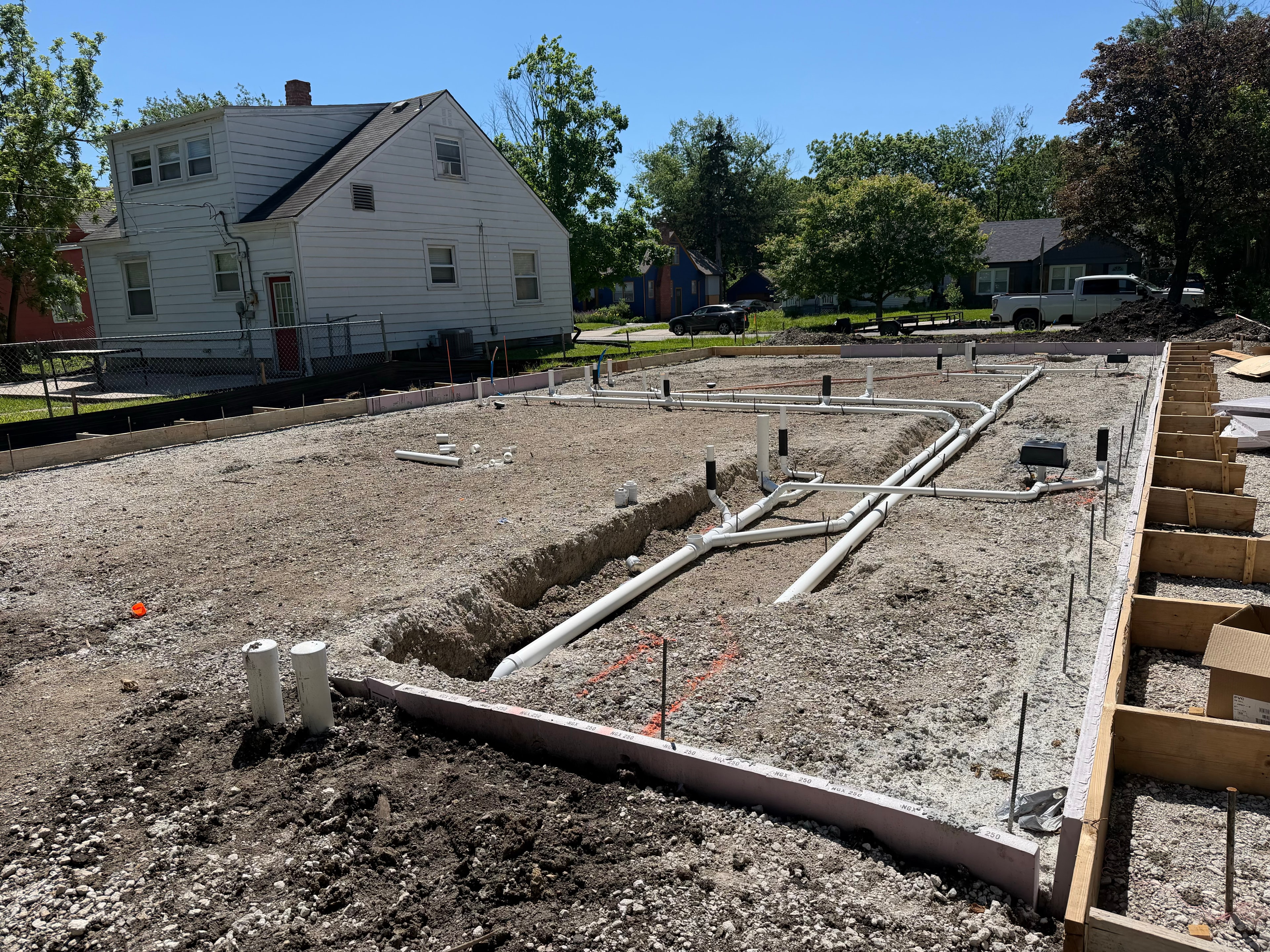 Underground plumbing roughed in across the full foundation footprint — PVC pipes trenched and stubbed up, foundation forms in place, blue Kansas sky overhead