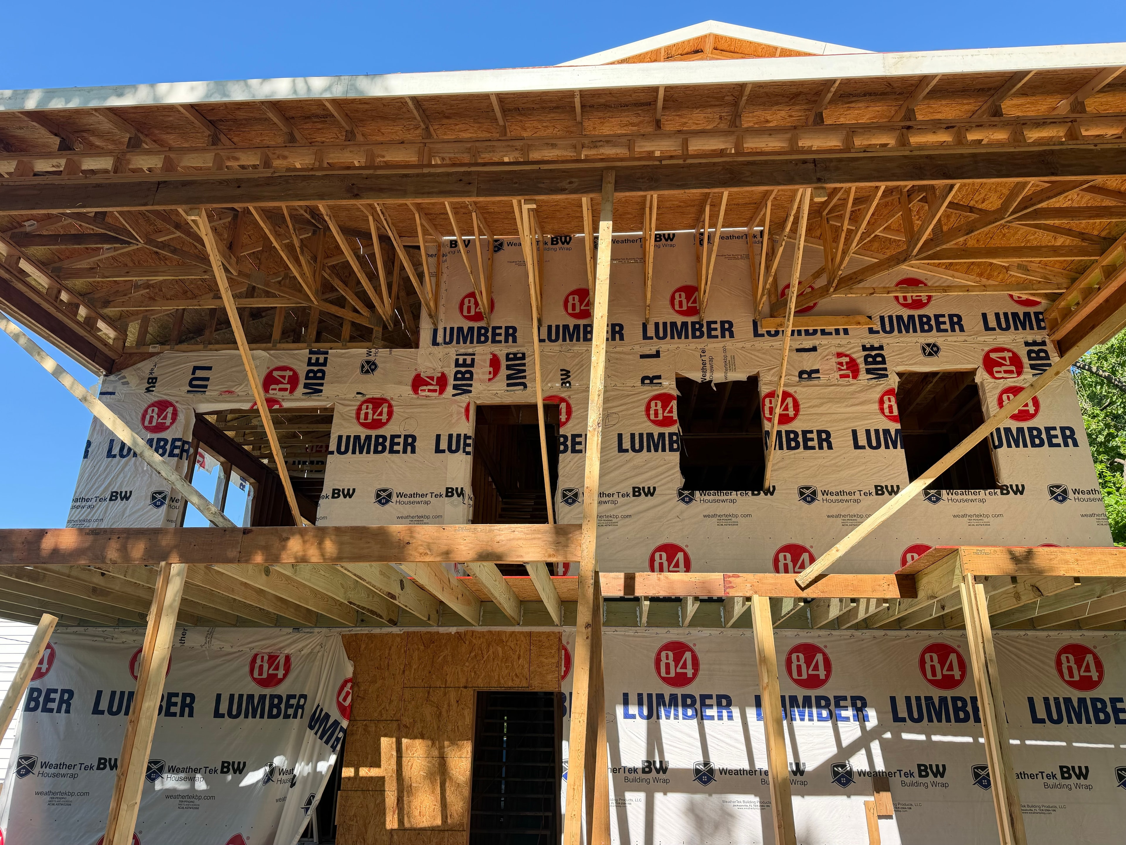 Looking up at two full floors of 84 Lumber house wrap — porch overhang framing in the foreground, window and door openings cut into the second floor, blue Kansas sky above the roofline