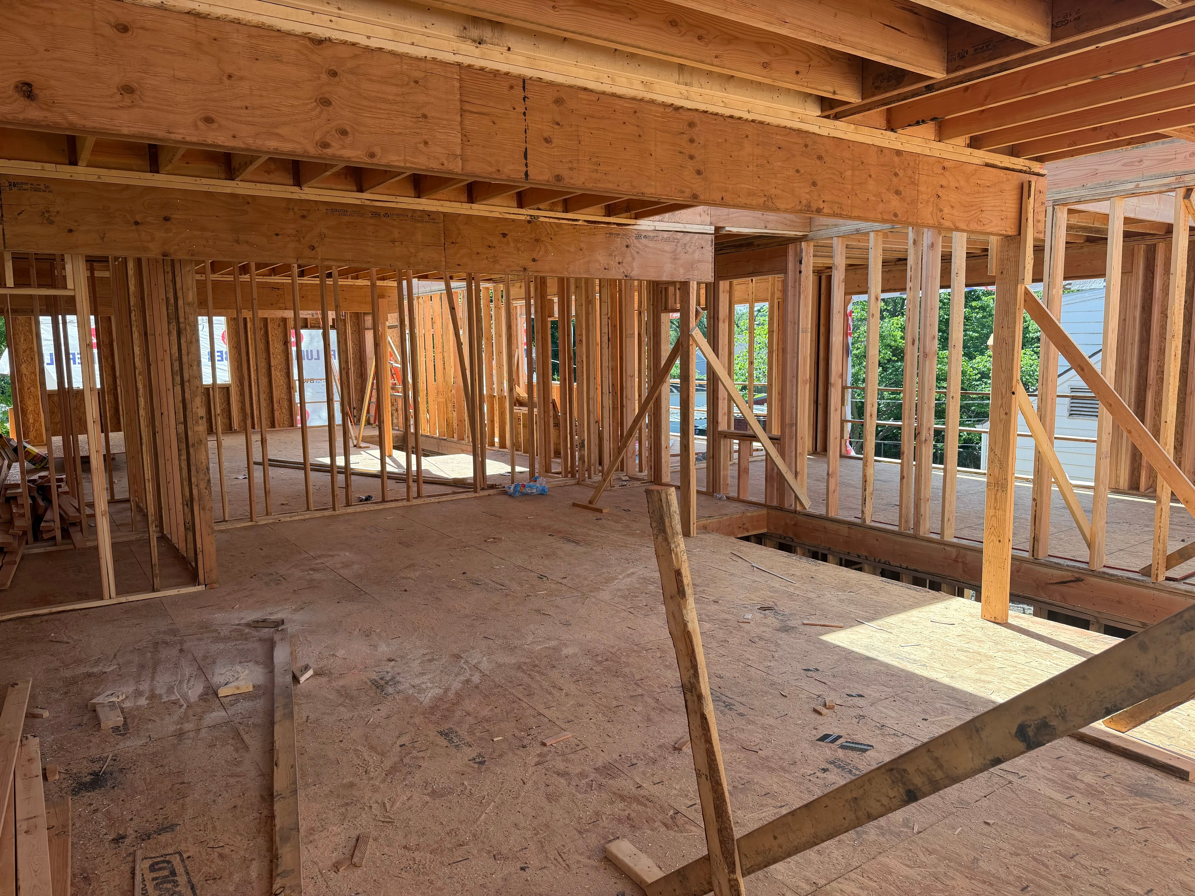 Standing inside the second floor — open framed rooms, ceiling joists overhead, stairwell opening in the foreground, neighborhood trees visible through the open walls