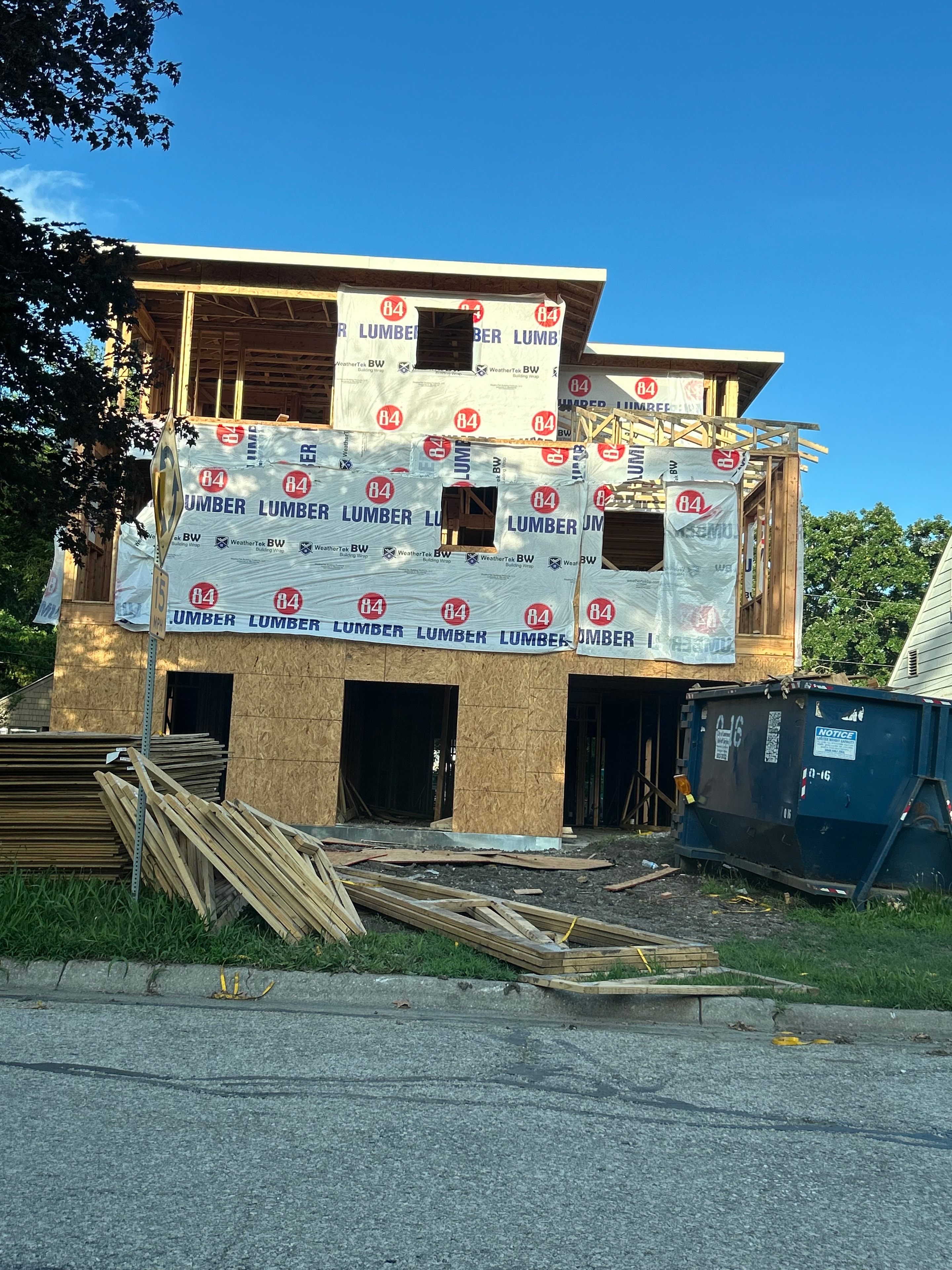 Street-level view of the front facade — 84 Lumber wrap covering both floors, window and door openings cut in, roof trusses visible at the top, lumber stacked to the right ready for the next phase