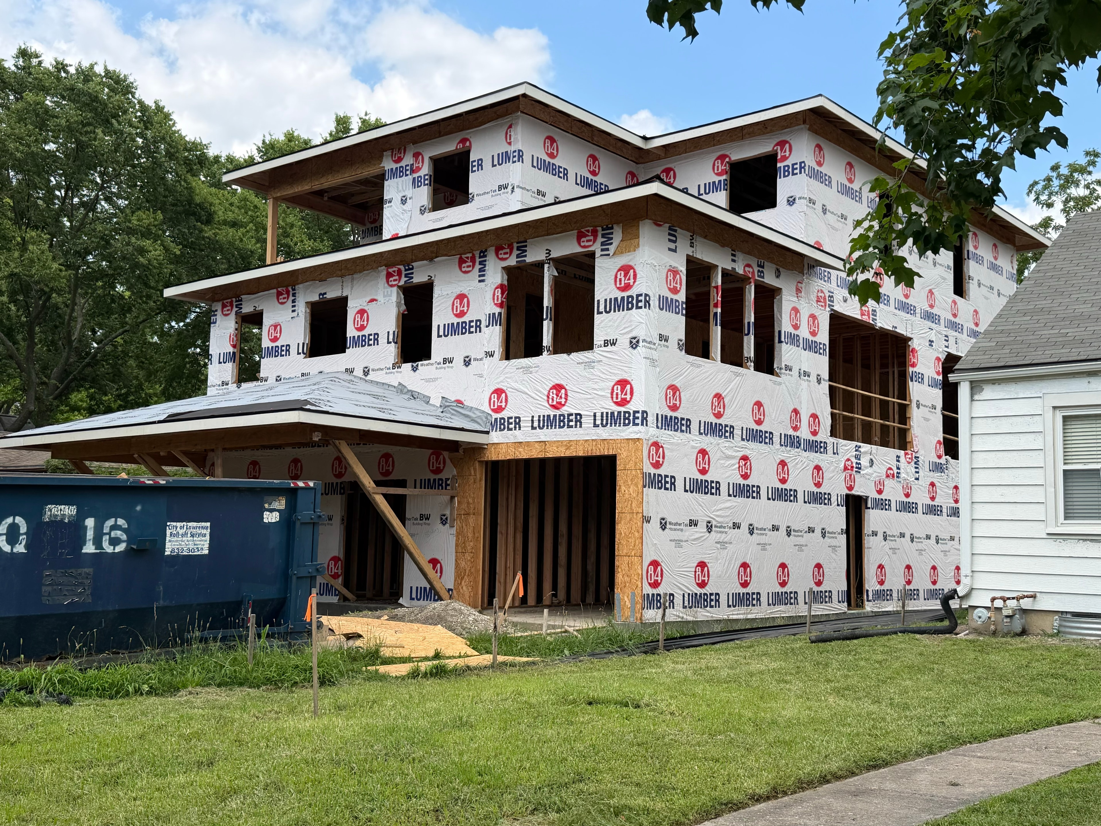 Corner angle of the full three-story structure — every inch wrapped in 84 Lumber house wrap, covered porch roof framed at ground level, neighboring house to the right showing the full scale of the build on the Lawrence lot. Look closely at the top right corner just past the tree leaves — that framed bedroom window on the third floor looks directly out at Lawrence High School's football stadium.