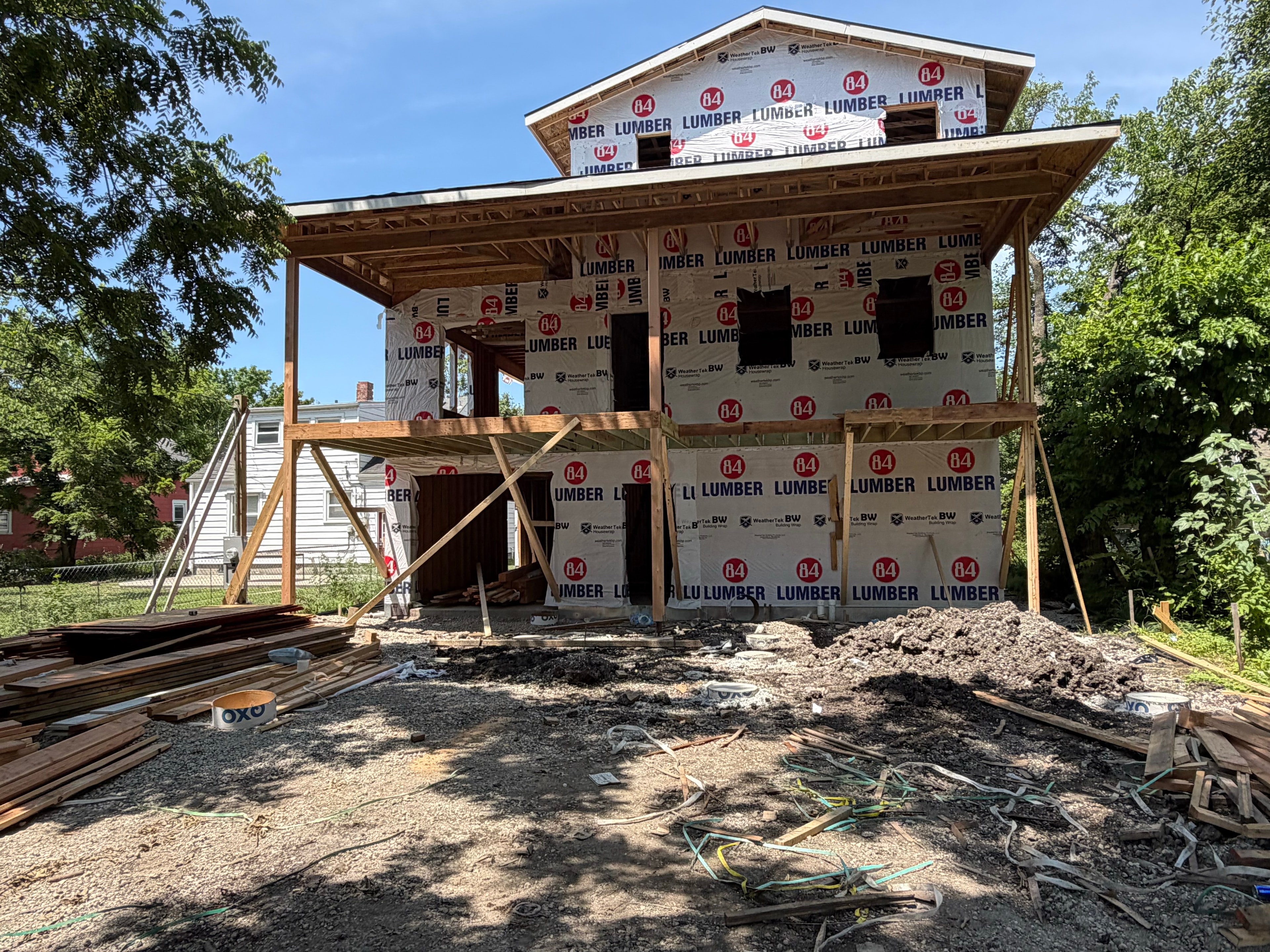Back view of the full three-story structure — 84 Lumber wrap covering every wall, covered porch deck framed on the second floor, window openings cut in, active work site with lumber and materials in the foreground