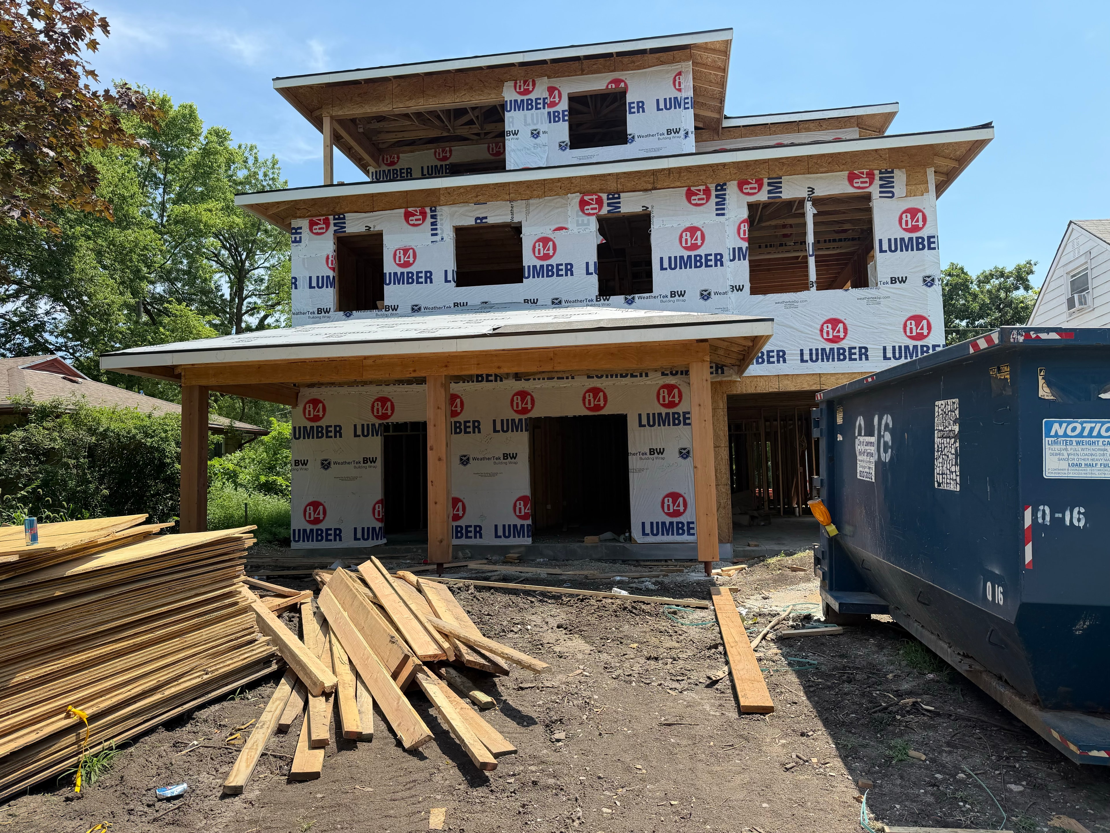 All three stories of 84 Lumber wrap on full display — covered porch framed at ground level, lumber stacked in the foreground, the entire build captured in one frame