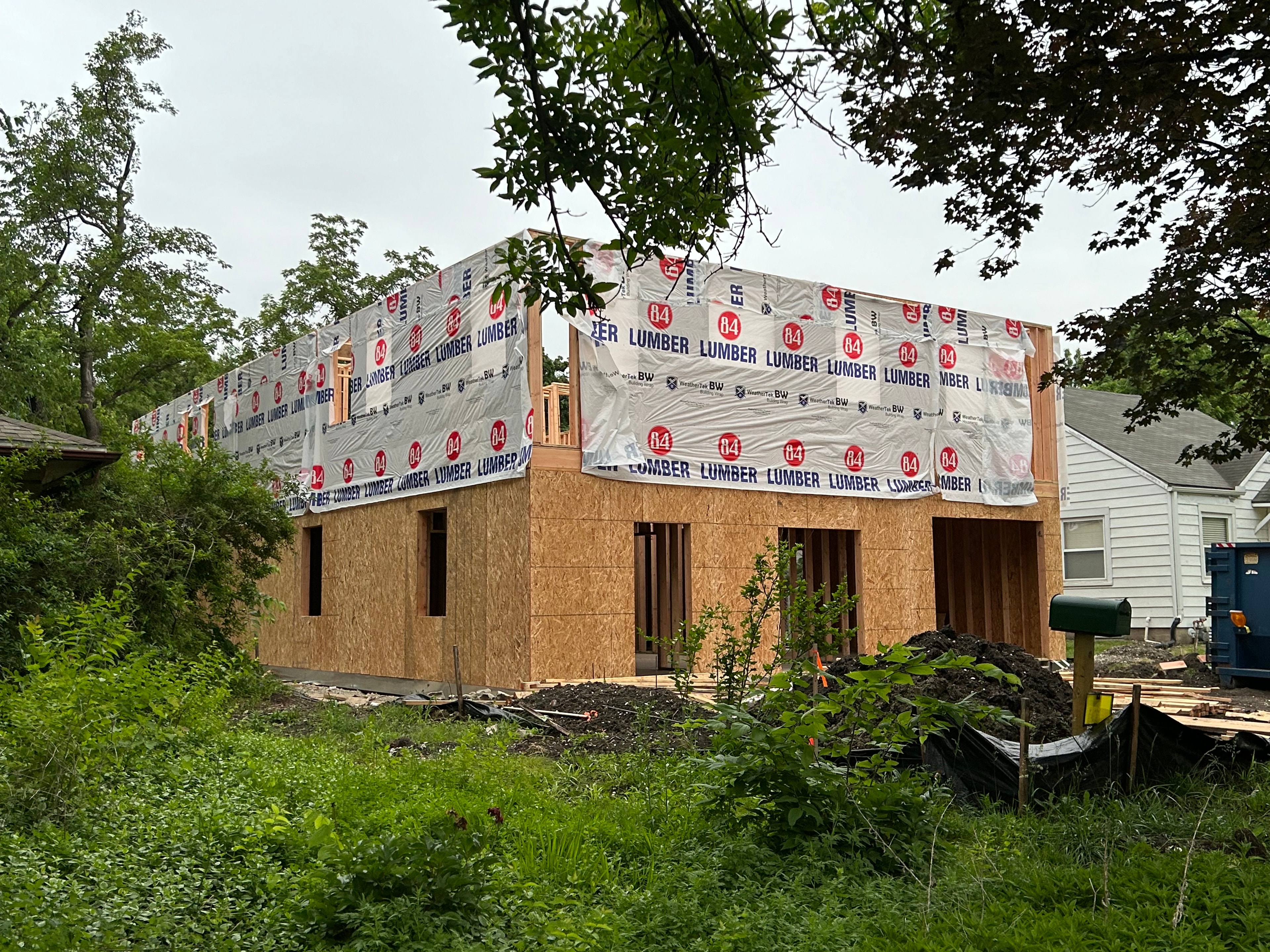 Aerial view through summer trees — first floor wrapped in 84 Lumber house wrap, second floor OSB walls rising with window openings framed out, the full footprint of the home visible from above