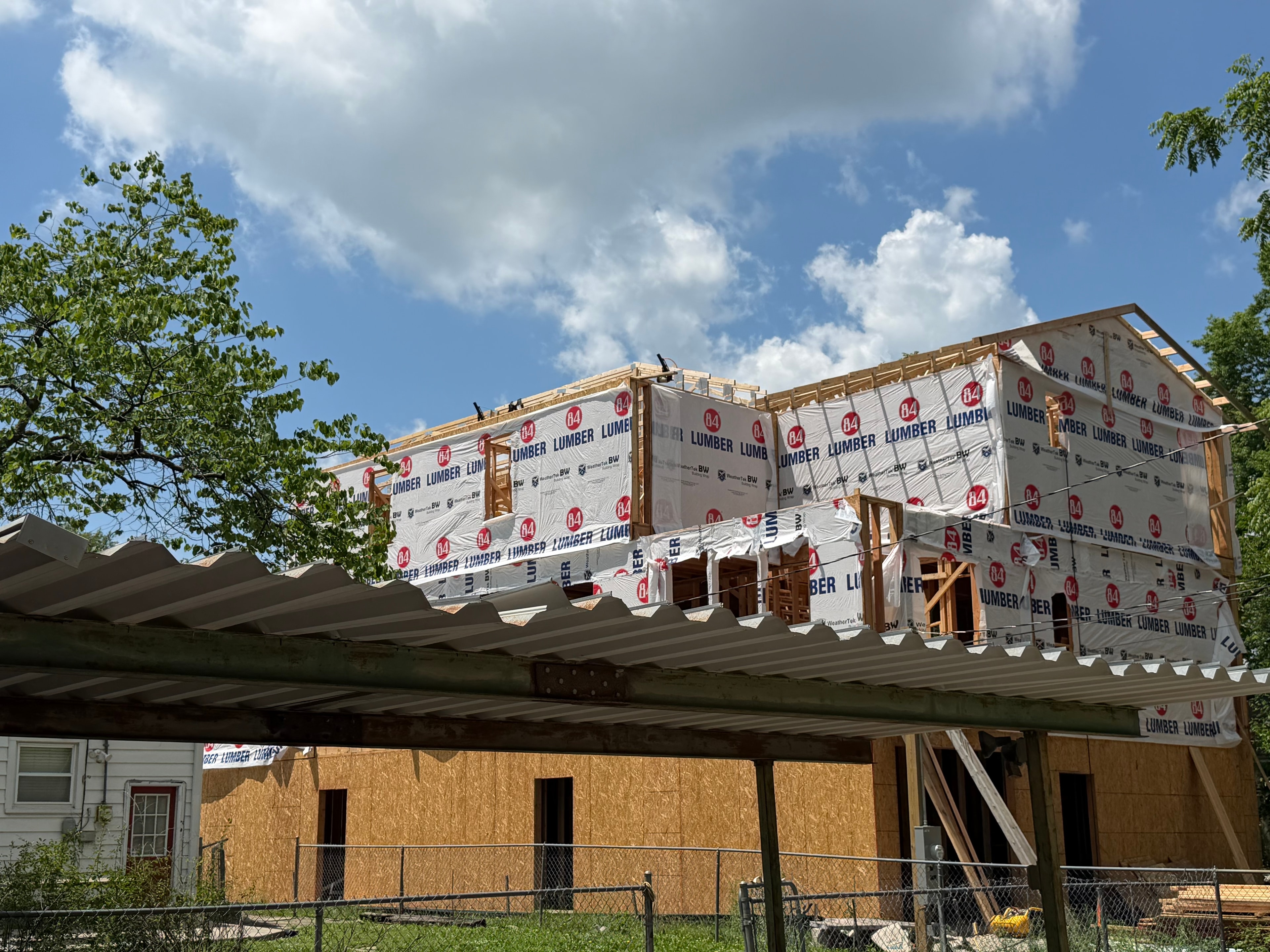 The full three-story structure wrapped in 84 Lumber house wrap rising above the neighboring roofline — framed against a brilliant blue Kansas summer sky